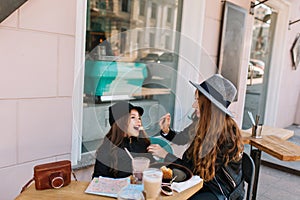 Amazing woman in retro hat having lunch with her daughter in black jacket, sitting at the table with camera and coffee