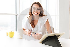 Amazing young lady sitting at the kitchen using tablet computer.