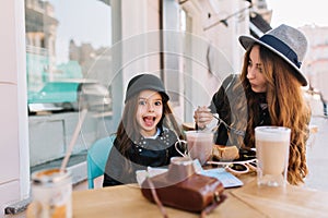 Amazing young woman in vintage gray hat eating tasty cheesecake with her little sister in weekend. Surprised brunette