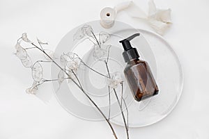 Amber glass cosmetic bottle. Shampoo pump dispenser on ceramic plate. Dry lunaria flower isolated on white table
