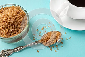 Amber sugar crystals in a spoon near a glass sugar bowl and a cup of hot drink over blue background. Unrefined brown cane sugar