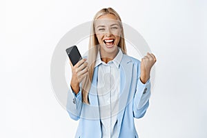 Ambitious businesswoman winning on mobile phone, cheering pleased and celebrating, standing in suit over white