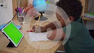 American African boy writing and talking in front of tablet screen at table in house interior.