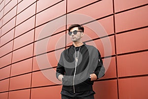 American attractive young hipster man with a fashionable hairstyle in stylish sunglasses in a vintage black jacket posing