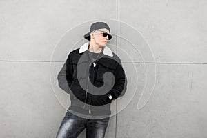 American attractive young man hipster in black fashionable jacket with white fur in dark sunglasses in a stylish baseball cap