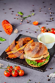 American burger with chicken cutlet, tomato, cucumber, green sauce. Sauce and for french fries on a black stone slate board