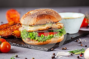 American burger with chicken cutlet, tomato, cucumber, green sauce. Sauce and for french fries on a black stone slate board