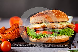 American burger with chicken cutlet, tomato, cucumber, green sauce. Sauce and for french fries on a black stone slate board