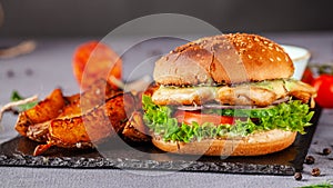 American burger with chicken cutlet, tomato, cucumber, green sauce. Sauce and for french fries on a black stone slate board