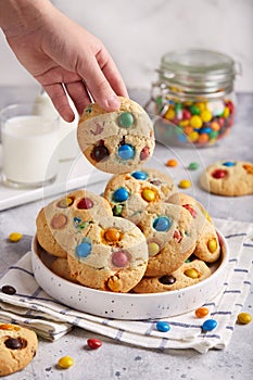 American cookies with colorful chocolate candy drops served with glass of milk. Child hand taking a cookie from a plate