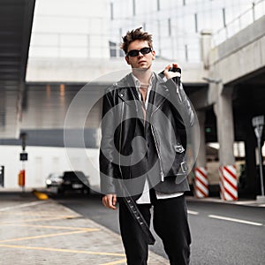 American fashion young man model in a stylish oversized leather black jacket in dark sunglasses in vintage jeans walks on the