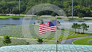 American flag waving over Sarasota National Cemetery with rows of white tomb stones on green grass. Memorial Day concept