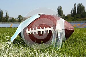 American football ball with hand sanitizer and protective mask on green field grass in stadium