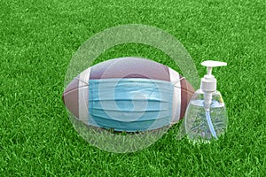 An American Football with a face mask and hand sanitizer on the right on field with green grass. Concept Football during pandemic