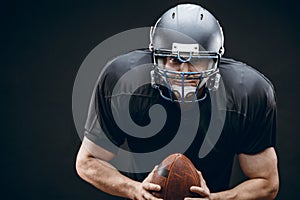 American football player in black sportwear with a ball on black background