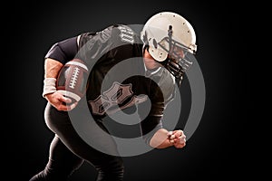 American football player in dark uniform with the ball is preparing to attack on a black background.