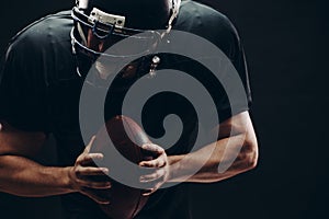 American football player with helmet and armour with a ball against black wall