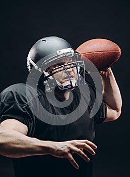 American football player with helmet and armour with a ball against black wall