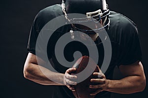 American football player with helmet and armour with a ball against black wall