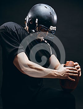 American football player with helmet and armour with a ball against black wall