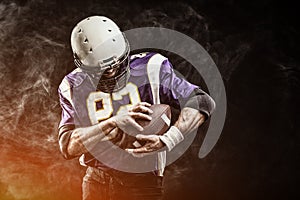 American football player holding ball in his hands in smoke. Black background, copy space. The concept of American