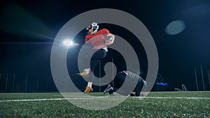 American football player jumps over another player, while competing on an empty field.