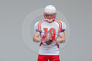 American football sportsman player wearing helmet holding rugby ball isolated on grey background