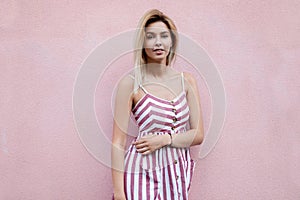 American glamorous young woman with blond hair in a stylish striped pink dress posing standing near a pink vintage building