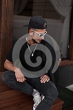 American hipster young man with a beard in a cap in a T-shirt in a jacket in sunglasses sits on wooden stairs near a vintage cafe