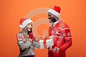 American man gifting Christmas present to happy girlfriend on orange background