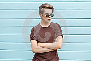 American modern hipster young man in stylish sunglasses in trendy brown t-shirt stands near a vintage wooden blue house