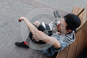 American modern young man in a trendy cap in a denim vest in stylish black ripped jeans in sneakers sits next to a hoverboard