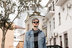 American nice young hipster man in a fashionable blue denim jacket in sunglasses stands on the street in the city. Handsome guy