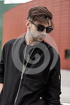 American nice young hipster man in a fashionable jacket in stylish sunglasses with trendy hairstyle walks in the city