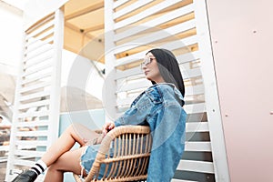 American pretty young woman model in fashionable sunglasses in stylish denim clothes sits on a vintage straw chair on the terrace
