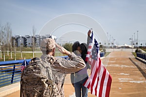 American soldier who has just arrived from a mission, saluting the American flag held by his wife who was waiting for him. Concept