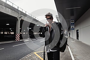 American trendy young man model in a youth oversized fashionable black leather jacket with a stylish hairstyle stands near modern