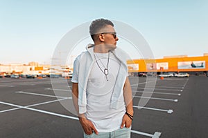 American urban young man in fashionable white summer clothes in trendy black sunglasses with a fashionable hairstyle stands on a