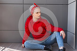 American young blond woman in black boots in a red bandana with a pattern in ripped jeans in a vintage red sweatshirt sitting