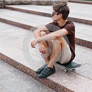 American young cool hipster man with hairstyle in stylish sunglasses in summer t-shirt in shorts in sneakers sitting