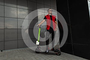 American young hipster man with a hairstyle in stylish denim red-black clothes in sunglasses stands with a modern electric scooter