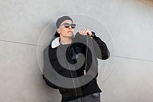 American young hipster man in stylish baseball cap in dark sunglasses in a fashionable black jacket with white fur in jeans