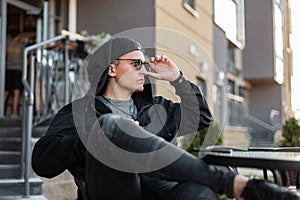 American young hipster man in stylish black clothes in fashionable sunglasses in a baseball cap in jeans sits in a summer cafe