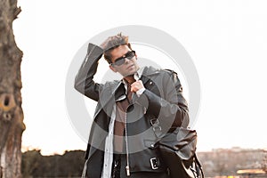 American young hipster man in stylish dark sunglasses in a trendy leather black jacket with a backpack posing near a wall in the