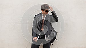 American young hipster man in stylish sunglasses in a trendy leather black jacket in jeans with a backpack posing near a wall in