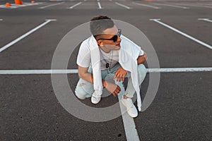 American young hipster man in sunglasses in blue jeans in white sneakers in a trendy T-shirt with a sweatshirt resting on a road.