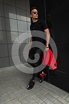 American young man in black fashionable clothes in stylish sunglasses with a vintage red jeans jacket in gym shoes relaxes