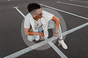 American young man model in fashionable sunglasses in vintage blue jeans in a stylish white t-shirt in sneakers sits on the