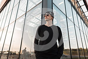 American young nice hipster man in vintage knitted black hat in stylish sunglasses in a t-shirt standing