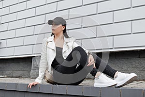 American young woman in a jacket in leather sneakers in black jeans in a stylish baseball cap with a backpack sits on a tile